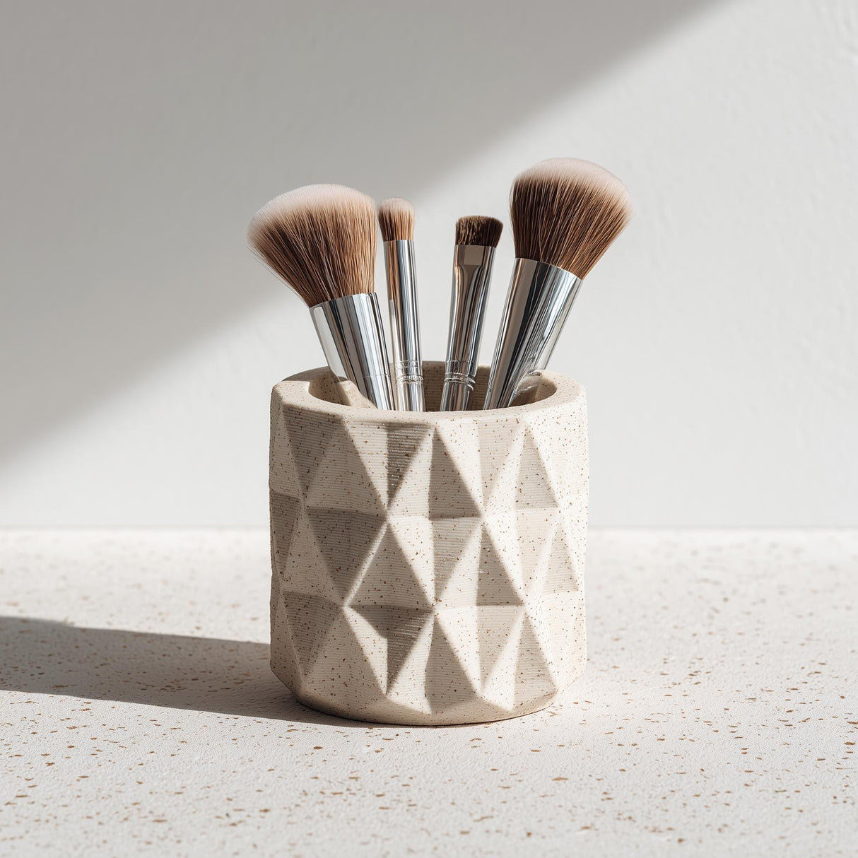 A beige 3D printed geometric cup holding makeup brushes, shown on a speckled surface with natural light.