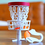 Collapsible mini disc golf basket on a desk with a disc and throwing tool for office trick shot challenges.