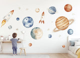 A child’s bedroom wall decorated with watercolor space-themed decals, including rockets, planets, and stars in blue.