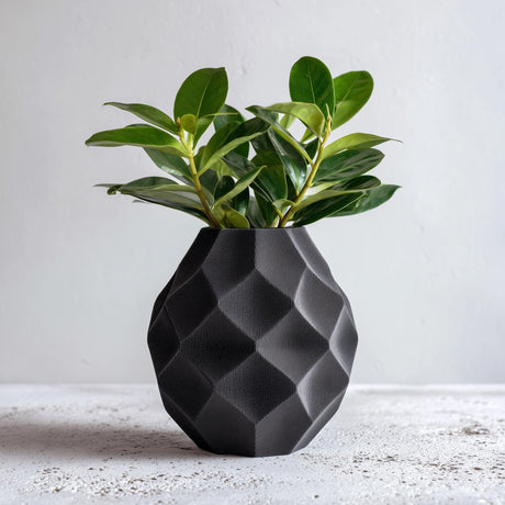 Black faceted 3D printed geometric vase holding green leafy stems on a white surface.