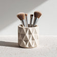 A beige 3D printed geometric cup holding makeup brushes, shown on a speckled surface with natural light.