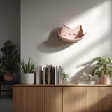 3D printed cat face wall shelf in soft pink, mounted above a wooden cabinet.