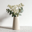 A tall, cream-colored 3D printed origami vase with vertical ribbed lines, holding white dried flowers and eucalyptus stems.