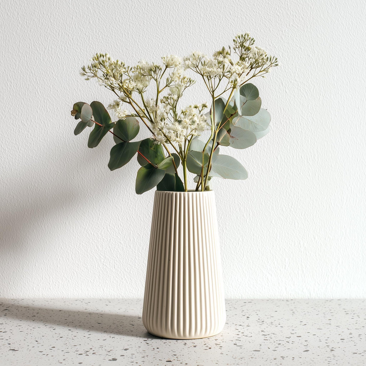 A tall, cream-colored 3D printed origami vase with vertical ribbed lines, holding white dried flowers and eucalyptus stems.