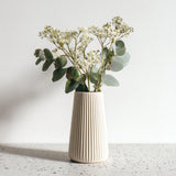 A tall, cream-colored 3D printed origami vase with vertical ribbed lines, holding white dried flowers and eucalyptus stems.
