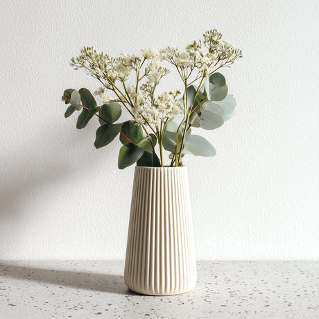 A tall, cream-colored 3D printed origami vase with vertical ribbed lines, holding white dried flowers and eucalyptus stems.