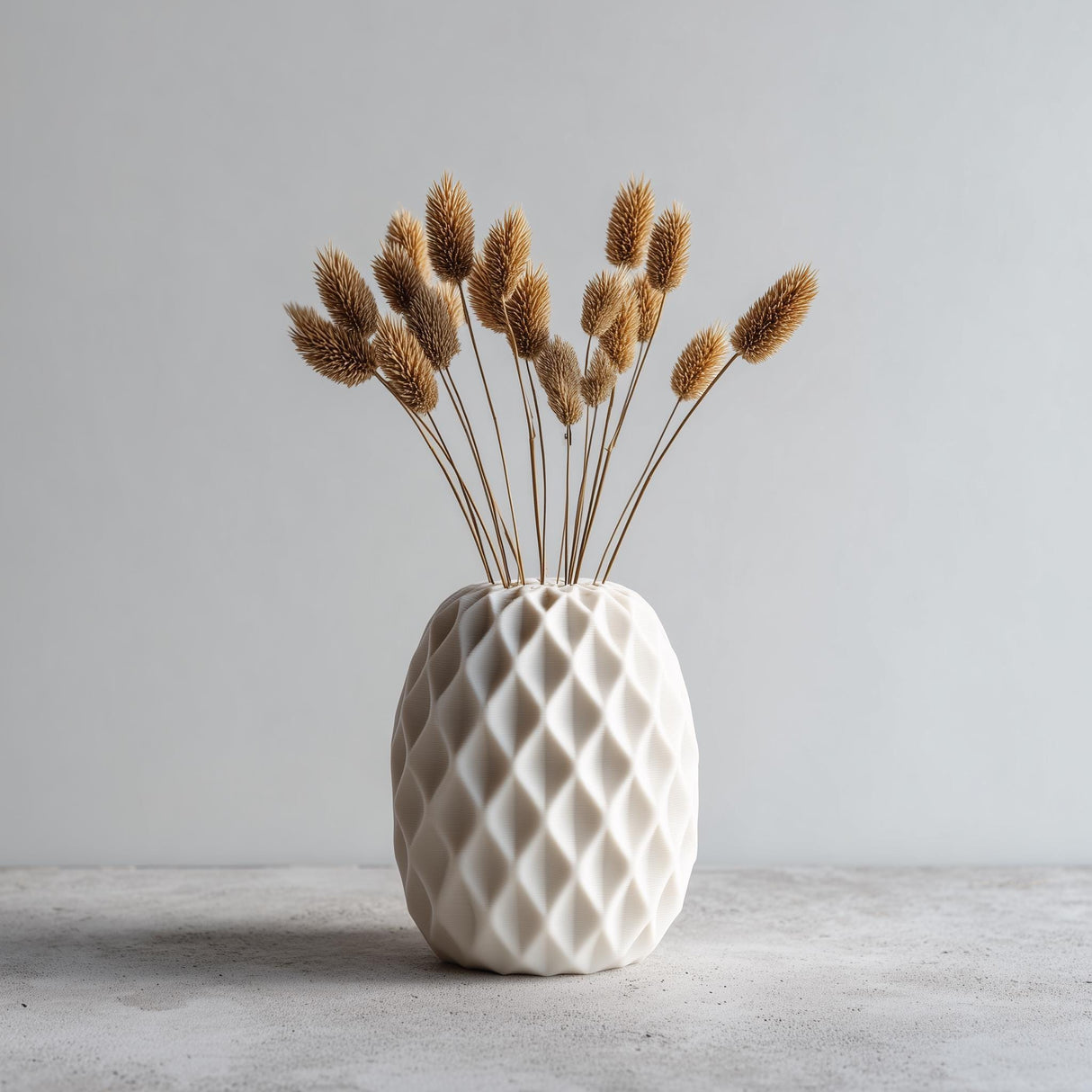 A white geometric 3D printed vase with a diamond-textured surface, holding dried beige flowers.