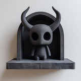 3D printed matte black wall shelf with a small horned spirit figure standing inside an arched niche, mounted on a light wall.