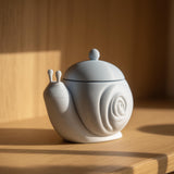 3D printed snail-shaped memorial urn with removable lid in matte PLA displayed on a wooden shelf with warm natural lighting.