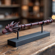 3D printed fantasy magic wand with dragon head handle and black display stand on a wooden table in warm ambient lighting.
