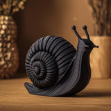3D printed snail sculpture in matte PLA with spiral shell texture displayed on a wooden shelf in warm cozy lighting.