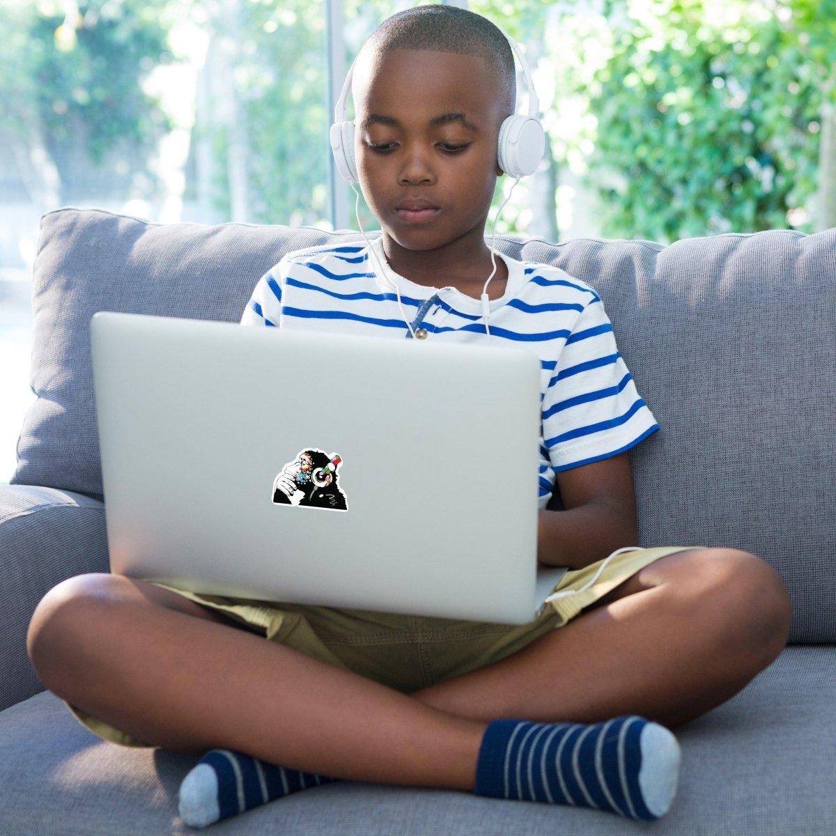 Child sitting on a couch with headphones, using a laptop featuring a contemplative monkey sticker.