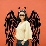 Woman posing with angel wings wall sticker, stylish outfit, sunglasses, against vibrant orange background.