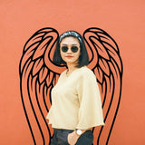 Woman in sunglasses standing against an orange wall with angel wings decoration in the background.
