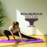 Woman stretching on a yoga mat with a workout decal saying 'Installing Muscles Please Wait' on the wall.