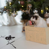 Siamese cat sitting in a festive box beside a Christmas tree and wall sticker design.